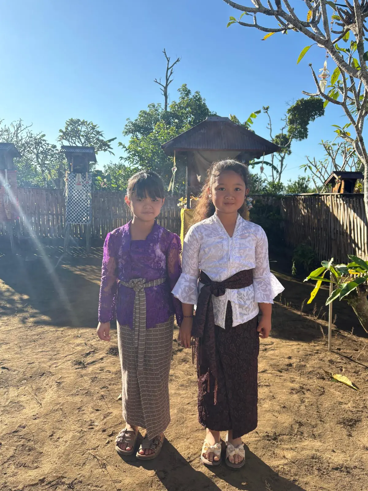 Two girls in Balinese temple dress standing together in the yard on a clear morning